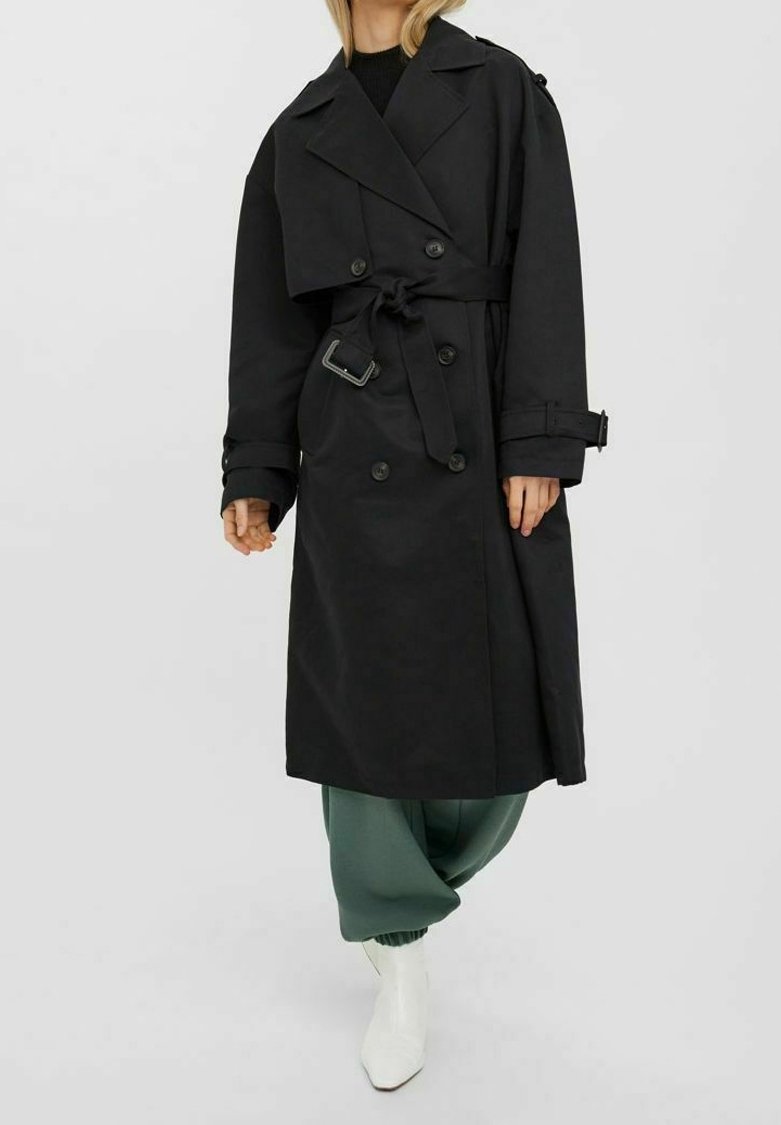 Czarny płaszcz trench z podwójnym zapięciem, szerokimi klapami, detalem w postaci pasa i zapinanymi mankietami, połączony z zielonymi spodniami i białymi butami.