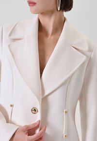 Blazer bianco doppiopetto in tessuto testurizzato, con rever a punta, dettagli con bottoni dorati e tasche laterali con bottoni a contrasto.