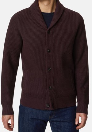 Homme portant un cardigan en tricot côtelé marron foncé avec des boutons et des poches à l'avant, par-dessus une chemise sombre et un jean bleu.