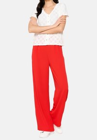 Top bianco a maniche corte con dettagli in eyelet e bottoni, abbinato a pantaloni rossi larghi e scarpe bianche.