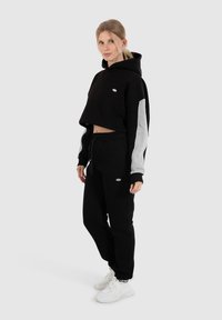 Zwarte cropped hoodie met grijze mouwen, voorzien van een klein logo. In combinatie met zwarte joggingbroeken en witte sneakers. Heeft een zachte stof en een relaxte pasvorm.