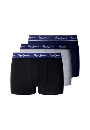 Junger Mann mit dunklem lockigem Haar, der dunkelblaue Boxershorts mit weißem Logo-Bund trägt, steht barfuß vor einem einfarbigen Hintergrund.