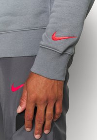 Felpa grigia con logo Nike rosso sulla manica, abbinata a pantaloni grigi con logo Nike rosa e un accento in rete nera.