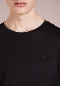 Schwarzes Baumwoll-T-Shirt mit Rundhalsausschnitt, ausgestattet mit einem dezenten, in dunklem Faden geprägten Logo auf der linken Seite der Brust.
