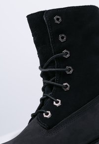 Schwarze Wildleder-Ankle-Boots mit sechseckigen Metallösen, runden Schnürsenkeln und einer strukturierten Sohle. Eine Steppnaht entlang der Basis bringt ein dezentes Design.