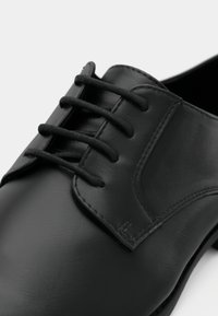 Chaussure habillée en cuir noir avec une finition lisse, un bout arrondi, un système de laçage avec des lacets noirs et des accents texturés sur la tige.