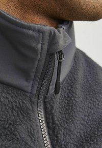 Grijze fleecJas met een hoge kraag, voorzien van een soepele stof en een gestructureerde fleece. Ritsluiting met zwarte hardware en een trekkoord.