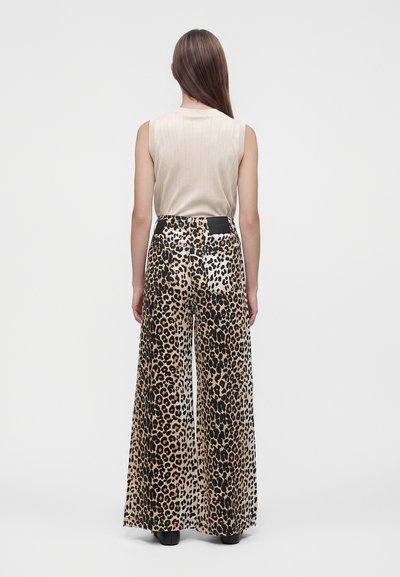 Personne debout, dos tourné, portant un haut beige sans manches et un pantalon large imprimé léopard avec des bottes noires sur fond uni.
