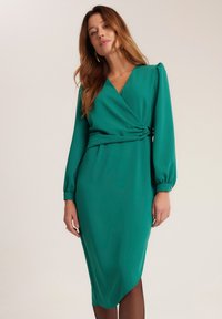 Zielona, do kolan sięgająca sukienka o fasonie kopertowym z długimi rękawami i marszczonym detalem w pasie. Gładki materiał o delikatnym połysku i miękkiej fakturze.
