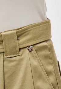 Χακί παν trousers με φαρδύ λάστιχο, πτυχώσεις και λεπτομέρεια από ασημένιο κουμπί λογότυπο. Το ύφασμα είναι μαλακό και με υφή.