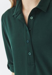Ciemnozielona koszulka polo z kołnierzykiem, trzema guzikami z przodu i podwiniętymi rękawami. Miękka tkanina o gładkiej fakturze.