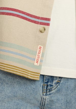Geweven shirt in beige met rode en blauwe strepen, voorzien van een knop en een label "REVOLUTION". Op de achtergrond staat een wit T-shirt en een spijkerbroek.