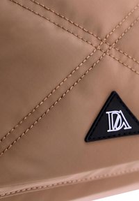 Tela de nailon color beige con costuras diagonales, que presenta un parche de logo triangular en negro. Textura suave con acentos en hilo beige en contraste.