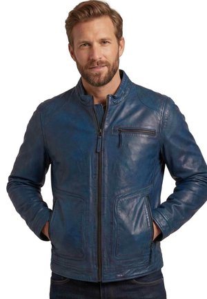 Mann mit hellbraunem Haar und Bart, der eine blaue Lederjacke mit Reißverschlusstaschen trägt, steht mit den Händen in den Jackentaschen.