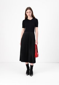 Abito midi plissettato nero con top aderente e maniche corte, abbinato a una borsa rossa. L'abito presenta un design strutturato e fluido.