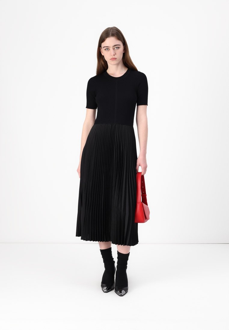 Robe midi noire plissée avec un top ajusté et des manches courtes, accompagnée d'un sac à main rouge. La robe présente un design texturé et fluide.