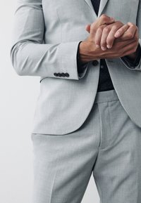 Giacca da completo grigio chiaro con tessuto strutturato e quattro bottoni scuri sui polsini, abbinata a pantaloni coordinati e una camicia scura con motivo sotto.