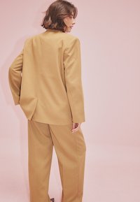 Woman with short brown hair wearing a loose beige suit, standing with one hand on her hip against a pale pink background.