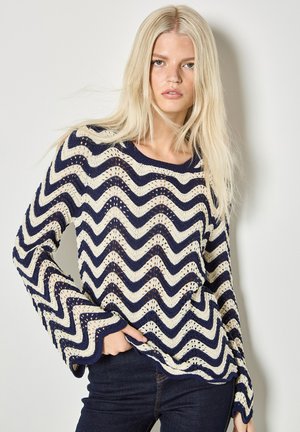 Femme blonde portant un pull en maille à manches longues à motifs zigzag bleu marine et blanc, associée à un jean foncé, posant devant un mur blanc.
