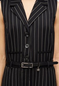 Blazer noir à fines rayures avec un col à revers, trois boutons noirs, deux poches avant et une ceinture noire ornée d'une boucle argentée et d'un charme.