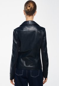 Giacca di pelle nera con colletto a punta e design aderente. Presenta una texture liscia e una silhouette su misura. Indossata con jeans scuri.