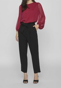 Donna che indossa una blusa trasparente a maniche lunghe di color bordeaux infilata in pantaloni neri a vita alta con cintura a sacco e tacchi aperti neri.