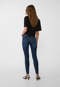 Camiseta corta negra combinada con vaqueros ajustados azules. Los vaqueros tienen bolsillos traseros y un ligero desvanecimiento. El modelo lleva sandalias de tacón negras.
