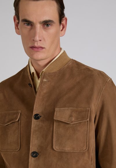 Jonge man met kort bruin haar, gekleed in een beige suède jas met voorzakken en zwarte knopen, neutrale lichtgrijze achtergrond.