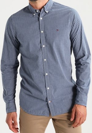 Muž v modrobílém kostkovaném košili s dlouhým rukávem a béžových kalhotách, zobrazen od krku po půlky stehen.