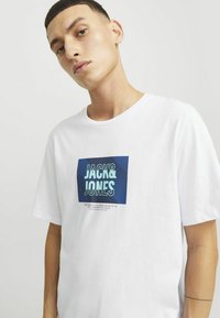 Valge puuvillane T-särk, mille esiküljel on sinine graafiline ristkülikukujuline logo koos tekstiga "JACK & JONES" ja peene triibulise mustriga.