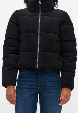 Personne portant une doudoune matelassée noire avec un col montant et une fermeture éclair argentée, associée à un jean en denim bleu.