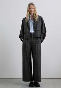 Traje de cuadros en tonos oscuros con una chaqueta corta y pantalones de pierna ancha. Superpuesto a una camisa de color azul claro, combinado con zapatos negros.