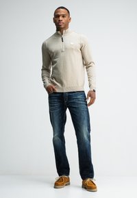 Beige rits-hals trui met lange mouwen, geribbelde manchetten en zoom, gedragen met donkerblauwe jeans en schoenen van bruin suède. Eenvoudig ontwerp, geen patronen.