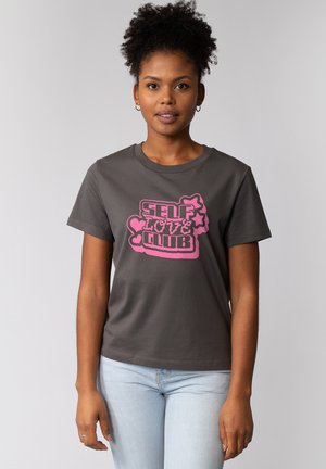 Frau mit lockigem Haar trägt ein graues T-Shirt mit rosa Aufdruck "SELF LOVE CLUB" und Formen, kombiniert mit hellblauen Jeans, steht vor einem grauen Hintergrund.