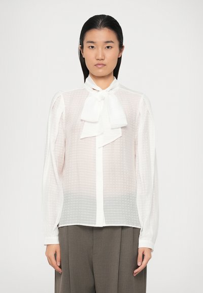 Blouse blanche texturée à manches longues avec un nœud au niveau du col, comportant des rayures verticales et une fermeture à boutons sur le devant.
