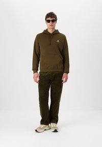 Sudadera verde oliva con cordón y bolsillo tipo canguro, combinada con pantalones de pana verde oscuro. Zapatillas marrones con detalles blancos. Gafas de sol negras.