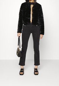 Svart faux fur-jacka med ribbat mönster, bärs över en ljus topp; kombineras med svarta jeans med kontrasterande sömmar och svarta sandaler.