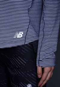 Granatowa koszulka w paski z długim rękawem z białym logo "NB". Charakteryzuje się eleganckim wzornictwem oraz czarnymi legginsami o teksturowanej powierzchni z geometrycznymi wzorami.