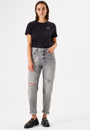 Zwarte katoenen t-shirt met een kleine dierenafbeelding, gecombineerd met grijze versleten jeans met gescheurde knieën en opgerolde boorden. Zwarte instapschoenen maken de look compleet.
