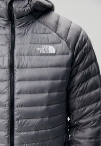 Jachetă puffer gri cu glugă, cu un model matlasat, fermoar frontal și logo în partea stângă sus a pieptului. Material neted, ușor.