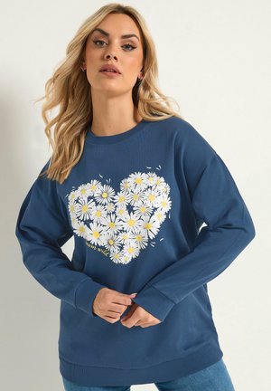 Femme blonde portant un sweat-shirt bleu avec un motif de marguerite en forme de cœur et le texte "rêve sauvage", posant devant un fond uni.
