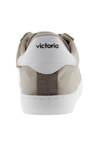 Sneaker beige avec un contrefort blanc, présentant un corps en tissu texturé et un grand logo de la marque en noir à l'arrière. Semelle en caoutchouc lisse.