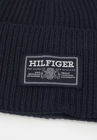 Berretto a coste blu navy con etichetta rettangolare nera e bianca con "HILFIGER" e il testo "GLI STATI DEL NEW ENGLAND."