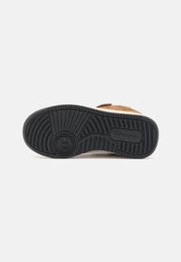 Suela de zapatilla con una parte inferior de goma negra y patrones de relieve texturizados para una mayor tracción, combinada con un detalle en ante marrón claro.