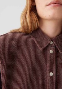 Close-up van een persoon met een bruine geribde corduroy shirt met een kraag en drie zilveren drukknoopjes.
