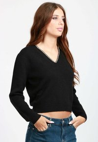 Una donna con lunghi capelli castani indossa un maglione corto nero a V e jeans blu, in piedi con le mani nelle tasche, guardando di lato.