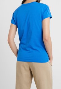 Modré bavlněné tričko s krátkými rukávy a kulatým výstřihem, zobrazené zezadu. Nošeno s béžovými volnými kalhotami.