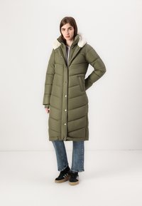 Pikk oliiviroheline polsterdatud mantel, millel on valeokka krae, pressitud õmblused, nupud ja küljetaskud, sobitatud teksade ja mustade spordijalanõudega.