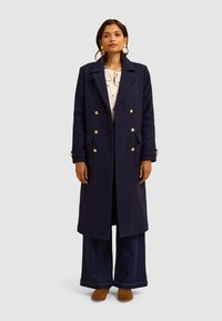 Cappotto blu in lana doppiopetto con bottoni dorati, tasche a toppa e silhouette dritta; abbinato a jeans in denim a gamba larga e scarpe marroni.