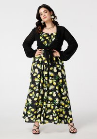 Een model poseert zelfverzekerd in een jurk met citroenpatroon en een zwarte cardigan, accessorized met statement oorbellen en ringen, tegen een witte achtergrond.
